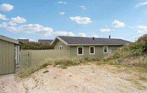 A single-story building with a sloped roof, a fenced yard, and a sandy ground.
