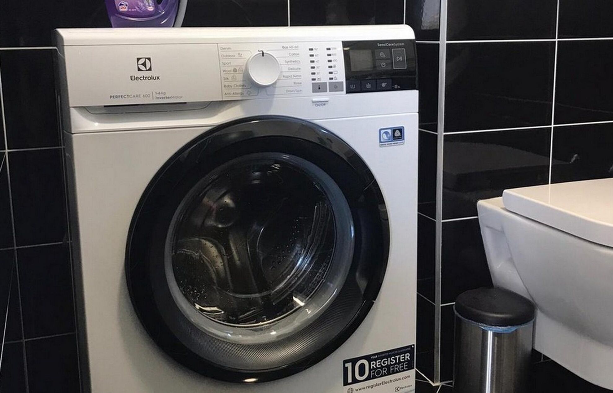 A front-loading washing machine with a digital control panel, a stainless steel trash can, and a white toilet in a bathroom with black tiled walls.