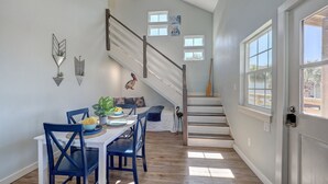 A dining area with a white table and blue chairs, a staircase with a wooden handrail, and a door with a view of the outside.