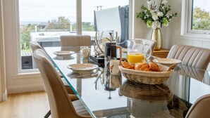 Ein Essbereich mit einem Glastisch, Stühlen, einer Kaffeemaschine, einem Orangensaftkrug, Croissants und einer Vase mit Blumen.