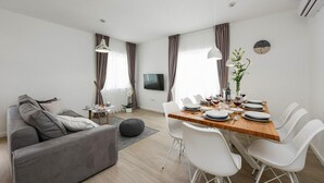 A modern living room with a grey sofa, a wooden dining table set for four, white chairs, and a TV mounted on the wall. - 3 bedroom lovely home in Nin (Nin)