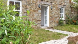 A stone house with a white door, a small porch, and a garden with green plants and flowers.