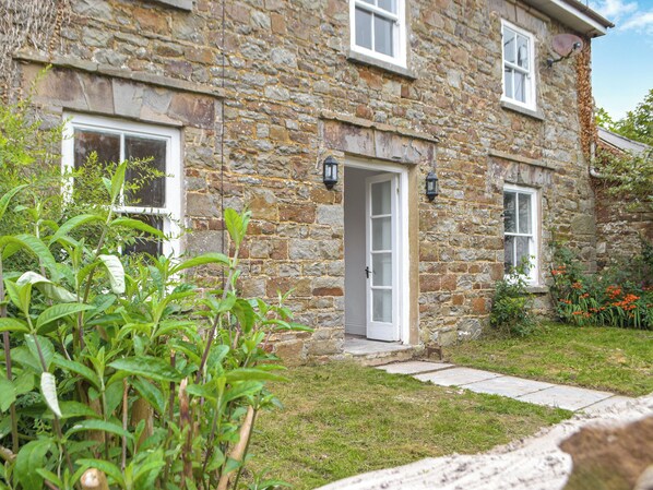 A stone house with a white door, a small porch, and a garden with green plants and flowers.