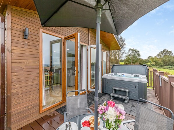 Une terrasse en bois avec un jacuzzi, des sièges d’extérieur et une table décorée de fleurs et de nourriture.