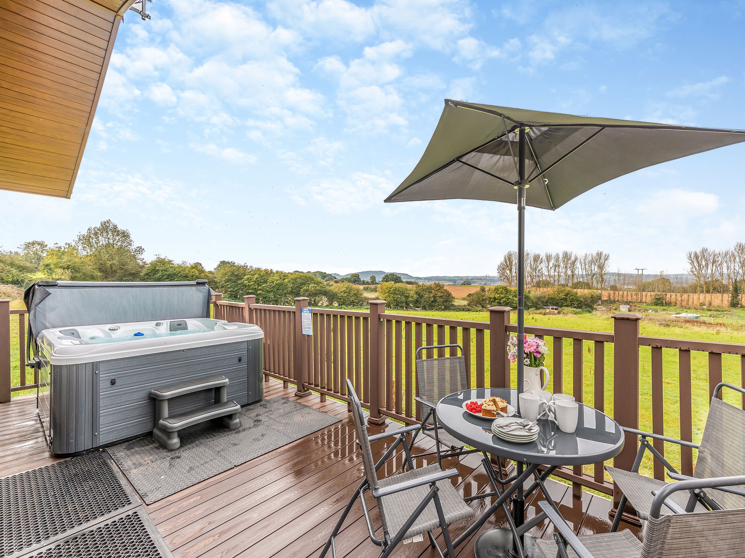Une terrasse en bois avec un jacuzzi, une table d’extérieur, des chaises et un grand parasol.