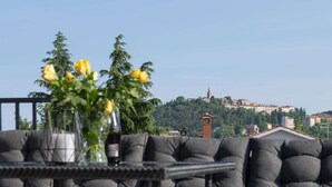 Eine Terrasse mit grauen Polstermöbeln und einem Tisch mit einer Vase voller gelber Blumen. Im Hintergrund ist eine Hügellandschaft mit Gebäuden und einem Turm zu sehen.