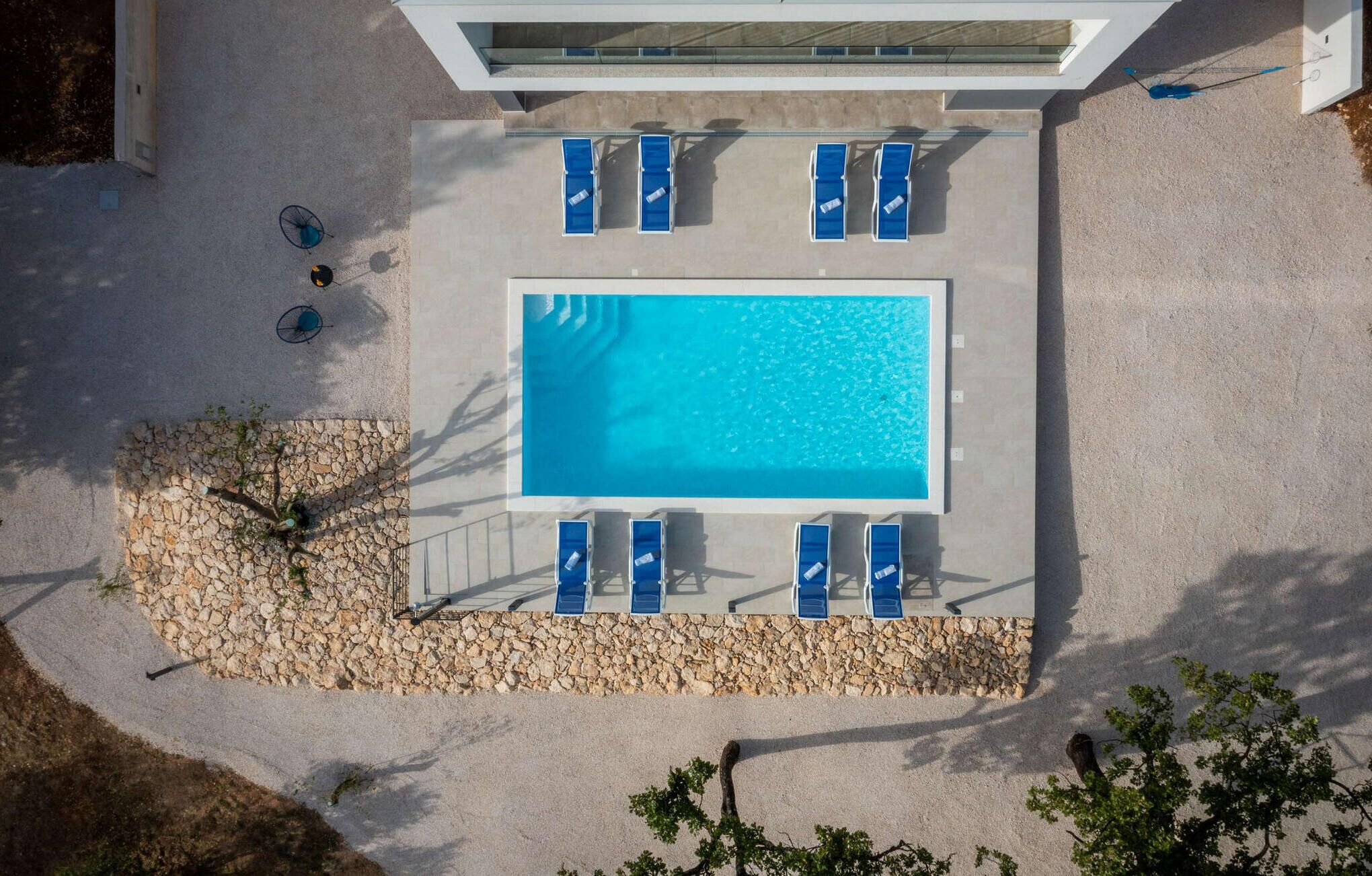 Eine Vogelperspektive auf einen Swimmingpool mit blauem Wasser, umgeben von einer Steinmauer und blauen Liegestühlen.