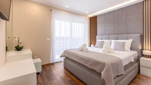 Un dormitorio moderno con una cama grande, una mesita de noche, un espejo y una ventana con cortinas translúcidas.