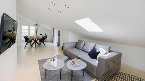 Un salón amplio con techo alto, tragaluces, un sofá gris y un comedor con mesa y sillas.