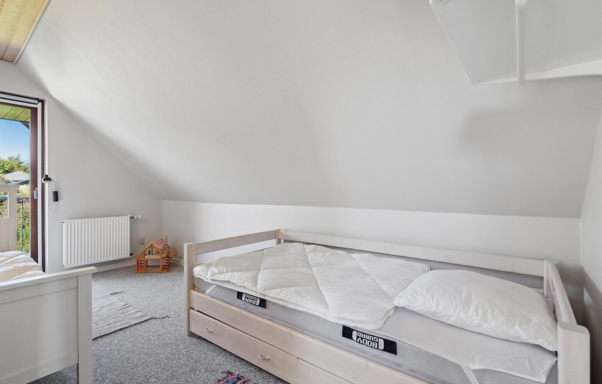 Una camera da letto con un letto singolo, una piccola casetta giocattolo in legno, un termosifone e una finestra vista verde.