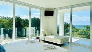 Ein modernes Schlafzimmer mit einem großen Bett, einer Couch und einem Fernseher. Große Fenster bieten Blick auf Grünflächen und das Meer.