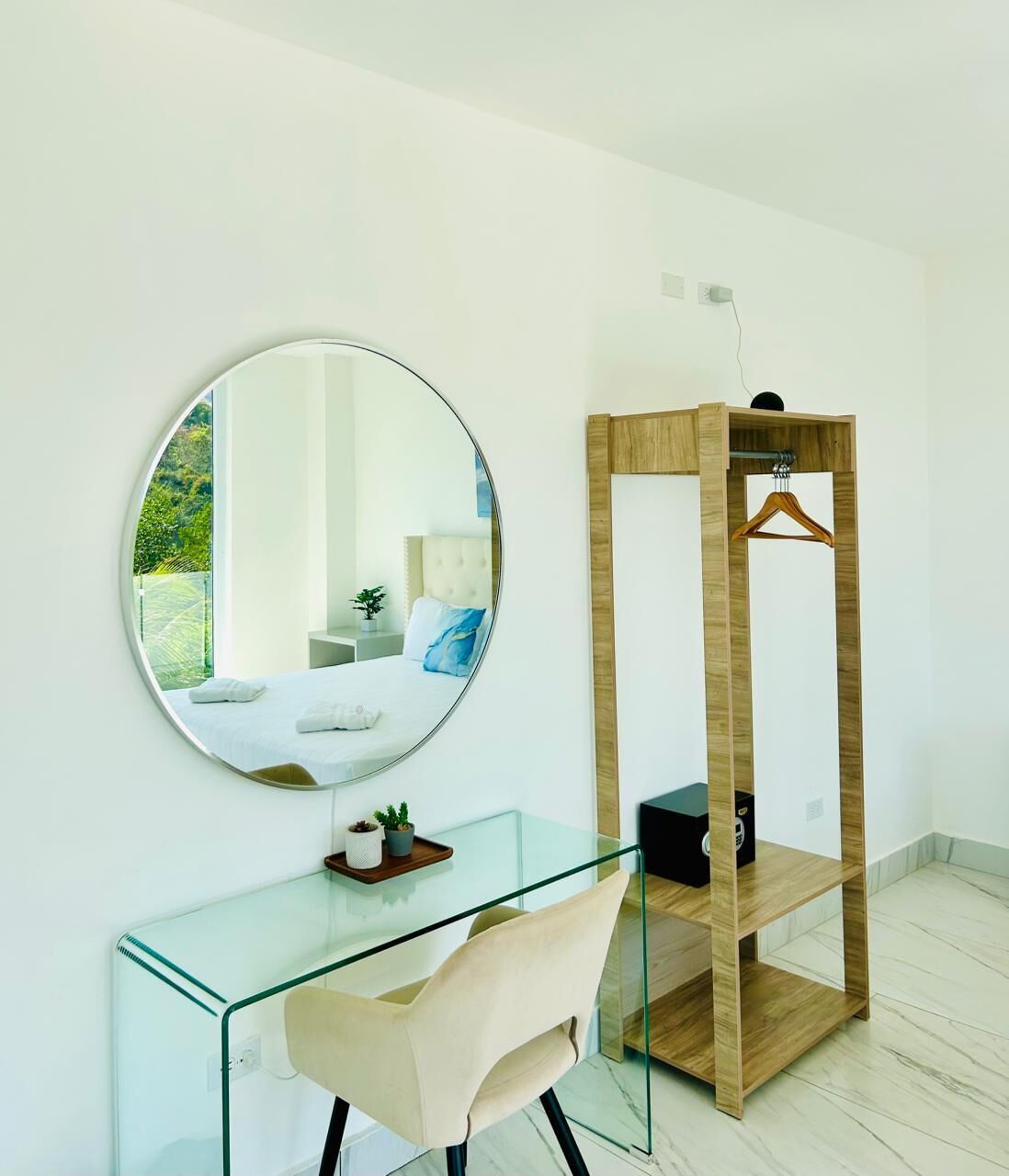 A modern room with a glass desk, a round mirror, a wooden hanger shelf, and a bed with a view.