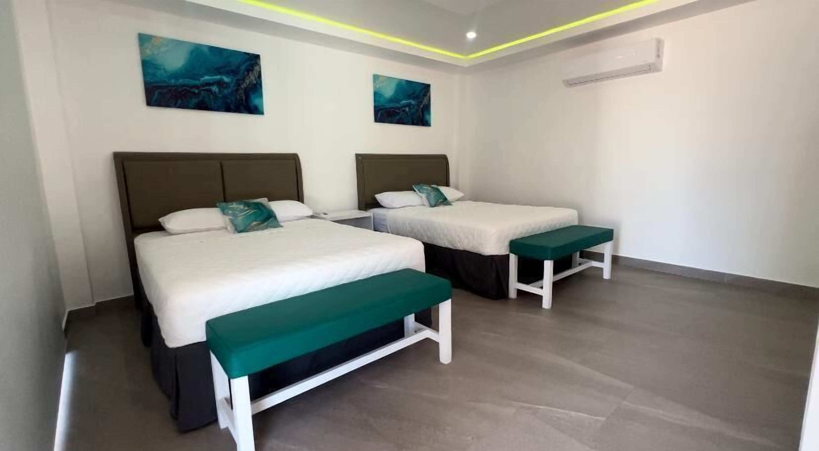 Habitación de hotel con dos camas, cada una con un banco verde, ropa de cama blanca y obras de arte azules en las paredes.