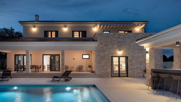 Una casa moderna de dos pisos con piscina, mobiliario de exterior y un muro de piedra.