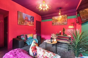 A vibrant room with a red wall, a sofa with colorful cushions, a small round table with a bottle and flowers, and a large potted plant. - Central Hull Stylish Home w/ Garden + Free Street Parking (Kingston upon Hull)