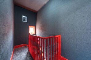 Een smalle trap met een rode leuning, een klein raam met zonlicht, en een structuurmuur.