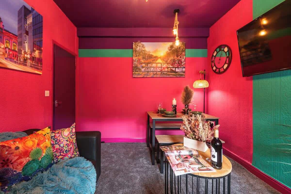 Ein Zimmer mit einer leuchtend roten Wand, einem blauen Sofa, einem Esstisch mit Stühlen und einem wandmontierten Fernseher.