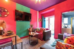 Ein lebendiges Wohnzimmer mit einem Flachbildfernseher, einem Esstisch gedeckt mit Tellern und Gläsern sowie einer gemütlichen Sitzecke mit einem farbenfrohen Sofa.