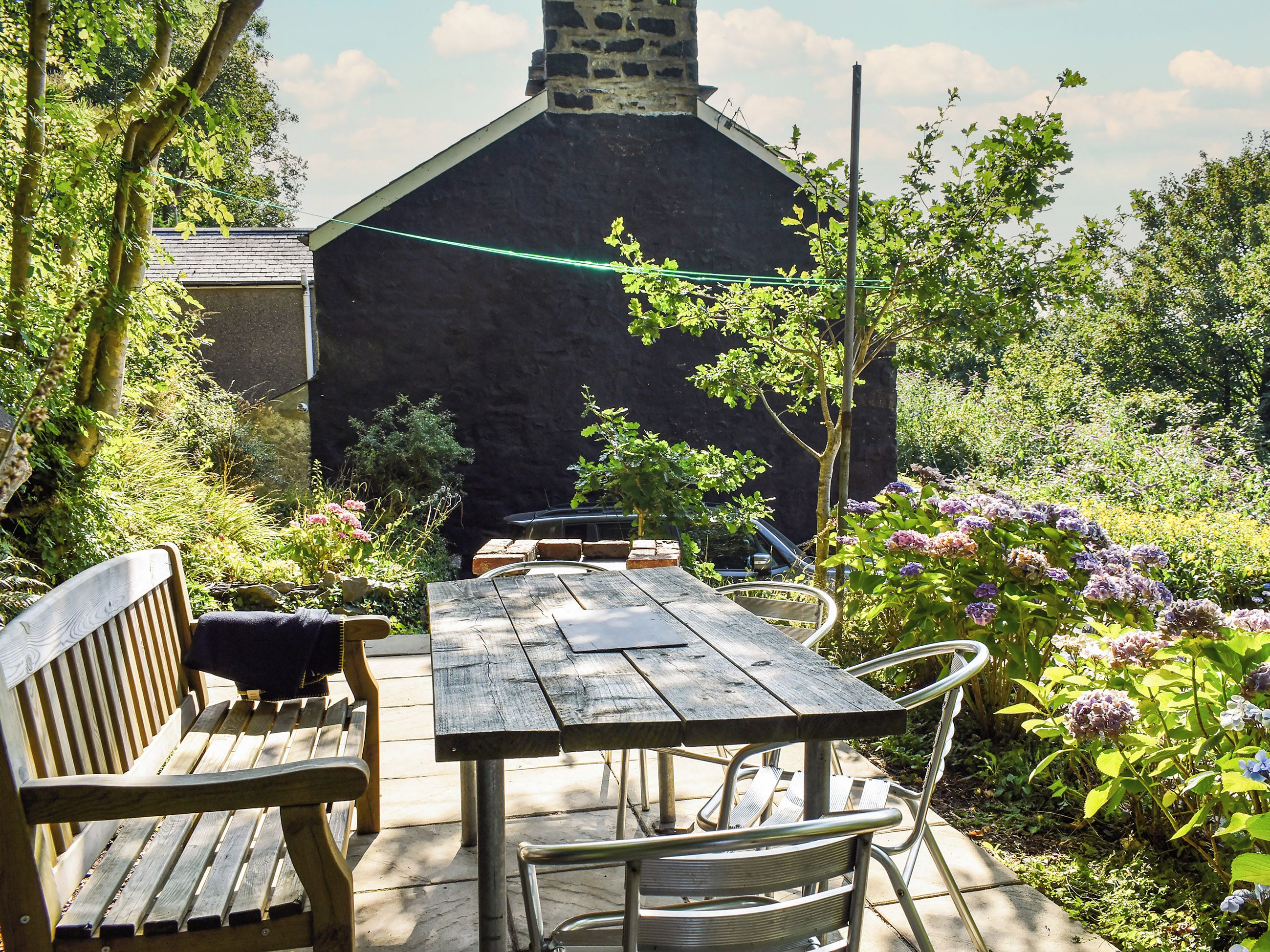 Une table et des chaises en bois, disposées à l’extérieur, devant un mur de pierre, avec un jardin en arrière-plan.