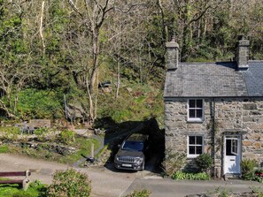 Une maison en pierre avec une voiture garée sous un pont, entourée d’arbres.