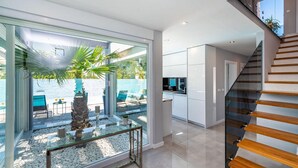 Ein modernes Interieur mit einer Glastür, die zu einem Poolbereich führt, einer Küche mit weißen Schränken und einer Holztreppe.