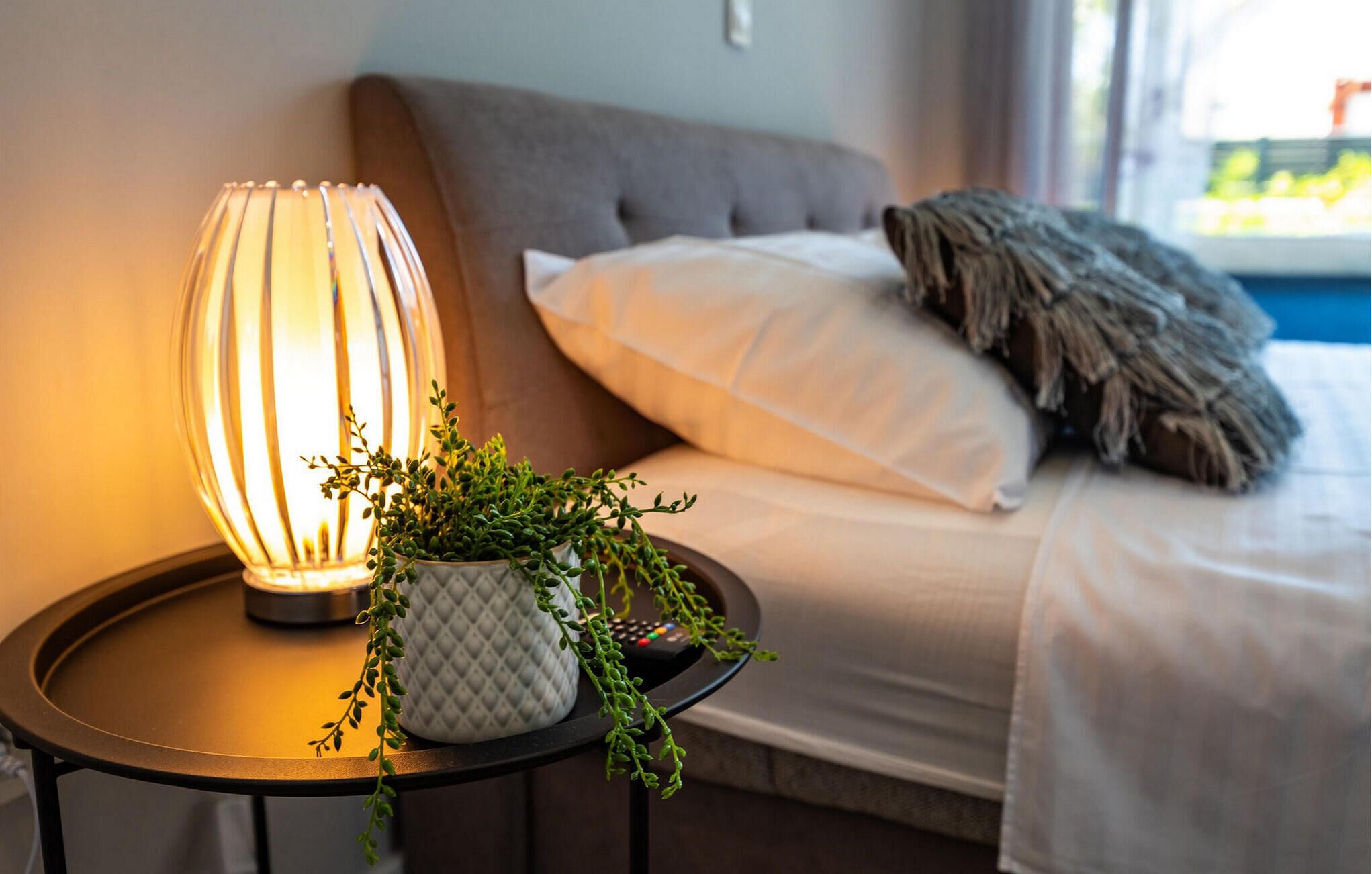 A neatly made bed with a white pillow and a grey throw blanket, a bedside table with a lamp and a potted plant, and a window with a view of greenery.