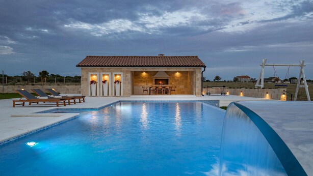 Ein Schwimmbecken mit einem Steinhaus im Hintergrund, eine Terrasse mit Liegestühlen und Blick auf die Landschaft.
