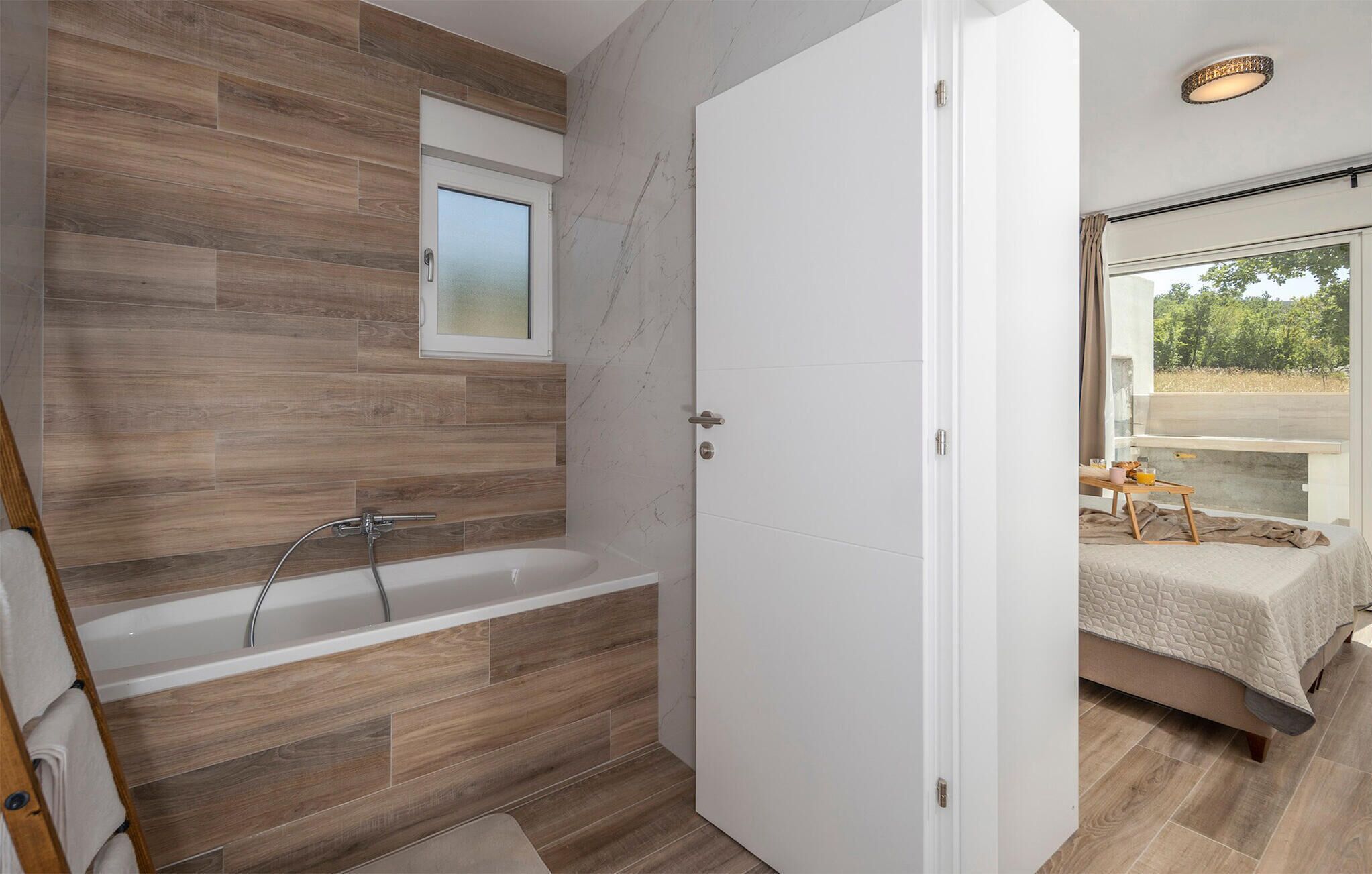A modern bathroom with a wooden floor, a glass shower enclosure, and a white door leading to a bedroom with a bed and a view of the outdoors.