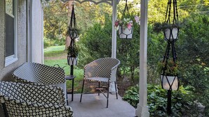 A covered patio with two patterned chairs, a hanging planter with flowers, and a view of a lush garden. - Hometract Cottage, Historic Property, UVA/Charlottesville/Ivy (Charlottesville)