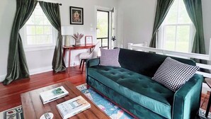 A living room with a green sofa, a coffee table, and a desk with a chair. There are two windows with green curtains, a framed picture on the wall, and a white door. - Hometract Cottage, Historic Property, UVA/Charlottesville/Ivy (Charlottesville)