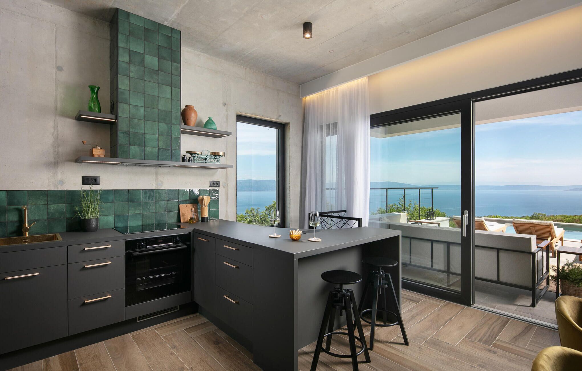 A modern kitchen with a large island, dark cabinetry, and a view of the sea through a large window.