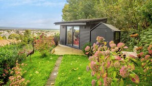 En lille, moderne hytte med en glasdør, omgivet af frodig vegetation og udsigt til en fjerntliggende by.