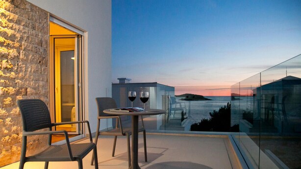Un balcon avec une table et des chaises en verre, deux verres à vin et une vue sur la mer au coucher du soleil.