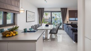 A modern kitchen with a dining area, featuring a central island with fruit, a dining table with chairs, and a sofa in the background. - Cozy home in Privlaka with sauna (Privlaka)