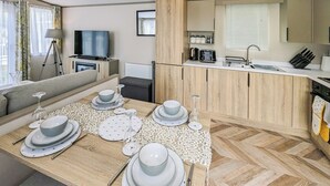 A modern kitchen with wooden cabinets, a dining area with a table set for four, and a living area with a sofa and a TV.