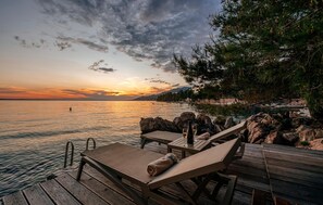 A wooden deck with lounge chairs and a table, overlooking a calm body of water with a sunset in the background. - Amazing home in Starigrad (Starigrad)
