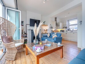 A modern living room with a glass coffee table, wicker chairs, a blue sofa, and a kitchen area with white cabinets and a large window.