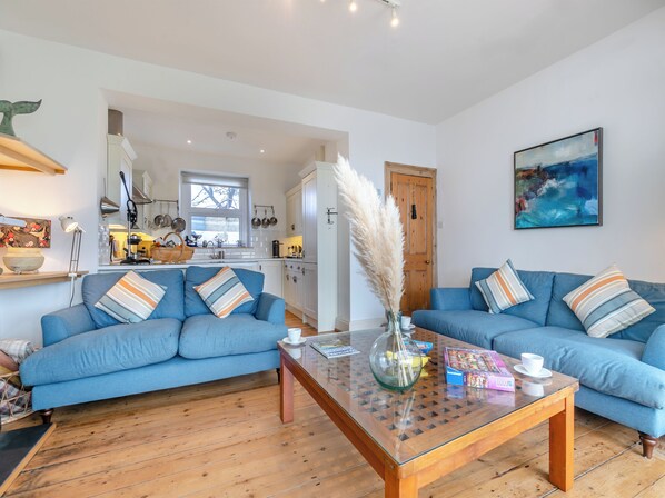 A modern living room with two blue sofas, a glass coffee table, and a kitchen in the background.