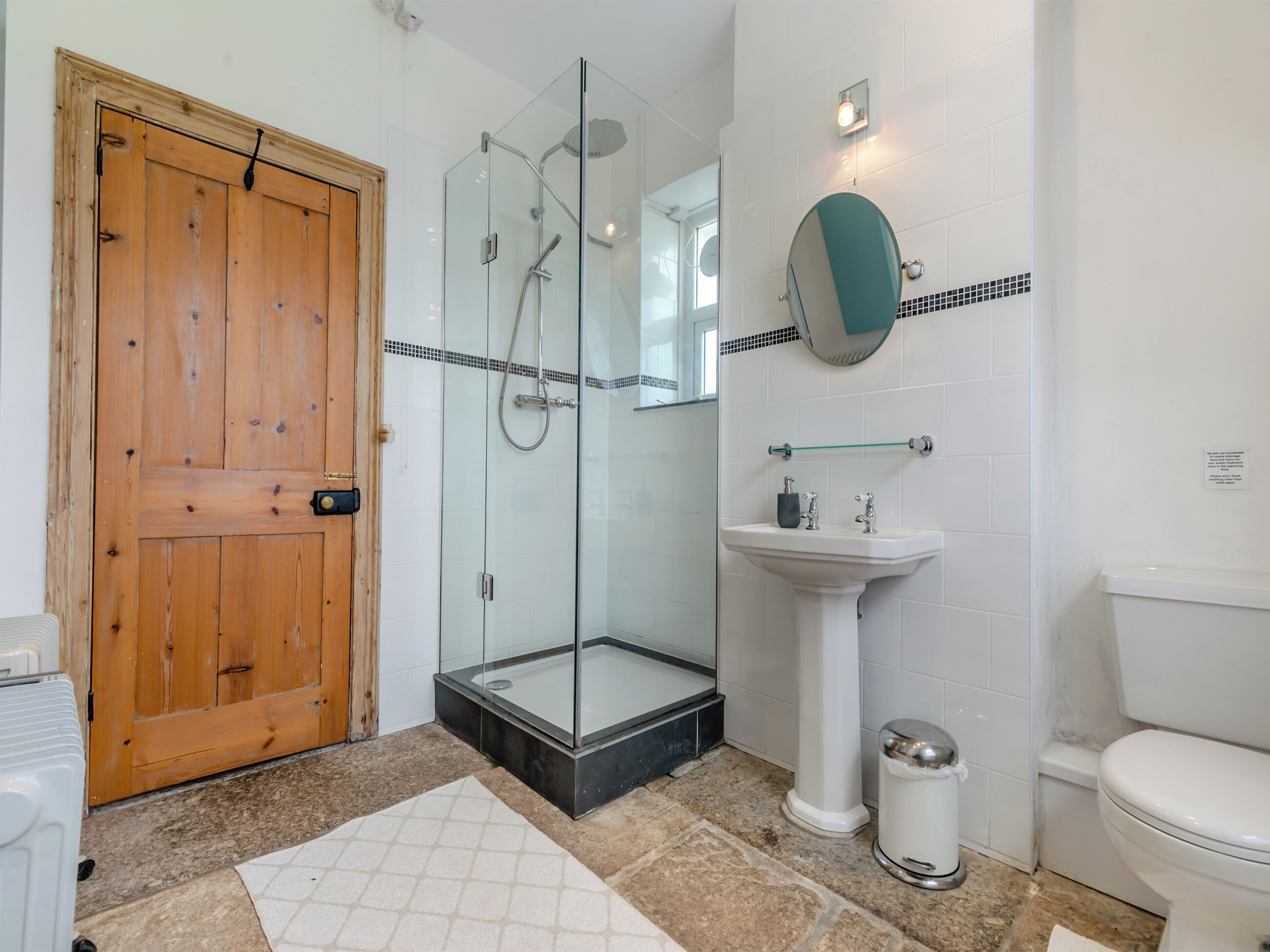 Un baño con puerta de madera, cabina de ducha de vidrio, lavabo sobre pedestal, espejo redondo y retrete blanco.