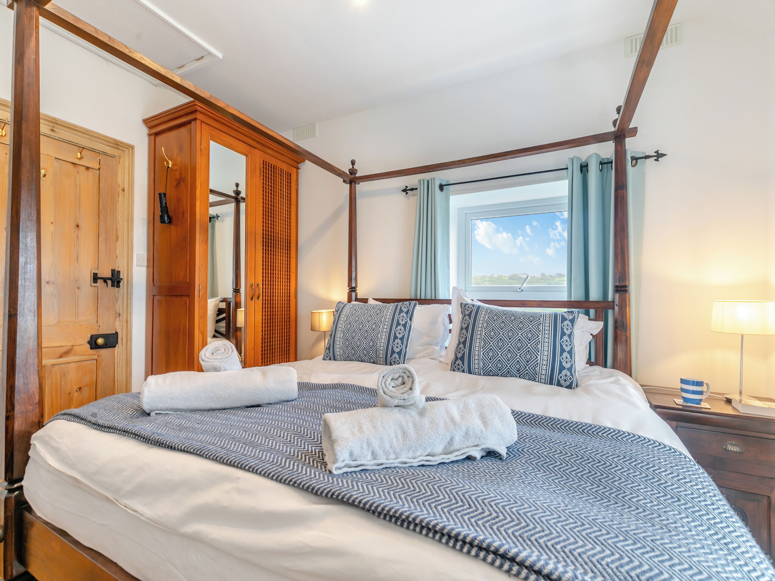 A four-poster bed with a patterned bedspread, a wooden wardrobe, a mirror, and a window with curtains.