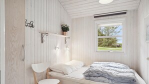 A small, single-bed room with a wooden slat wall, a window with a view of trees, a white chair, and a bedside table with a lamp. - Cozy home in Hjørring with WiFi (Hjørring)