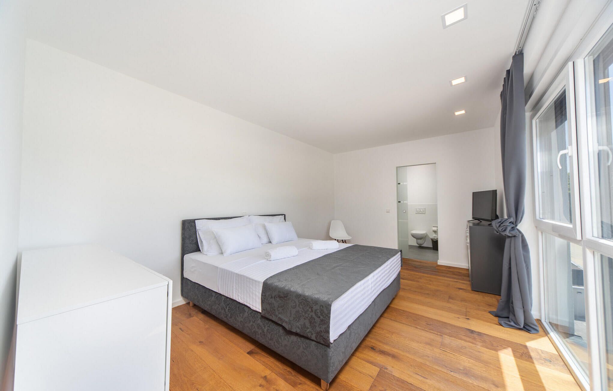 A modern bedroom with a large bed, a bedside table, a TV, and a bathroom visible through an open door.