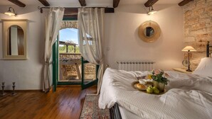 Ein Schlafzimmer mit einem Bett, einem Heizkörper, einem Fenster mit Vorhängen, einem Spiegel und Blick auf eine Steinsäule und Grünfläche draußen.