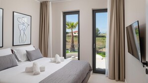 A modern hotel room with a large bed, a flat-screen TV, and a balcony with a view of palm trees and a red-roofed building. - Beautiful home in Nin (Nin)