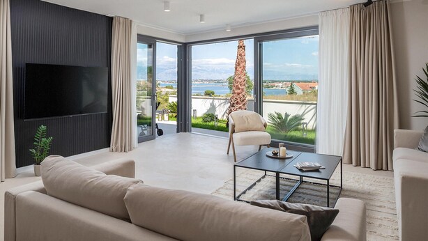 A modern living room with a large flat-screen TV, a sofa, a coffee table, and a chair. The room has large windows with a view of a body of water and greenery. - Beautiful home in Nin (Nin)