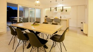 A modern dining area with a wooden table and black chairs, a kitchen with white cabinets, and a glass door leading outside. - Luxurious home in Zaton (Zaton)
