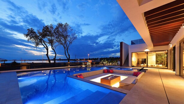 Un espace piscine extérieur moderne avec vue sur l’eau et les arbres, un coin salon couvert avec des coussins, et un escalier menant à un niveau supérieur.