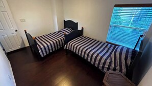 Una habitación pequeña y funcional con dos camas individuales, piso de madera, una ventana con persianas y una puerta.