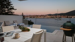 Um terraço com mesa, cadeiras, churrasqueira e vista para o horizonte urbano à beira-mar ao entardecer.