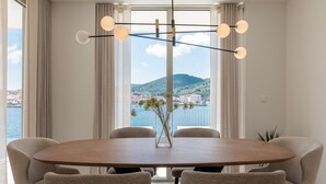 Uma sala de jantar com mesa redonda, seis cadeiras, um vaso com flores e vista para uma cidade costeira através de grandes janelas.